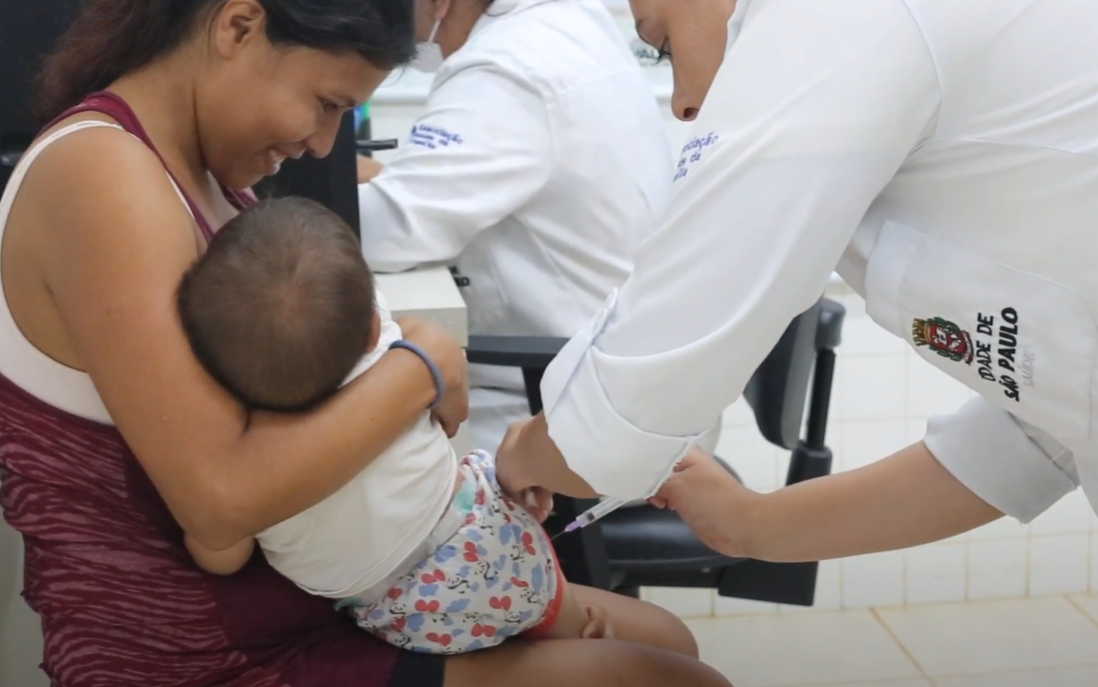 A foto mostra Amanda, uma mulher indígena de cabelos castanhos, lisos e presos, segurando seu filho no colo, Jonathan, de 7 meses, enquanto ele é vacinado na perna. Amanda está sorrindo